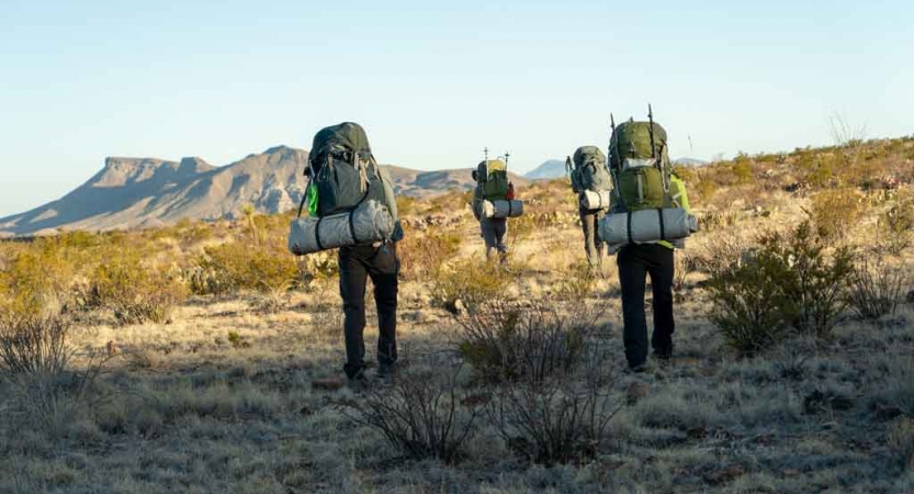 a group of gap year students carrying backpacks hike across a desert landscape toward a mountain in the distance  